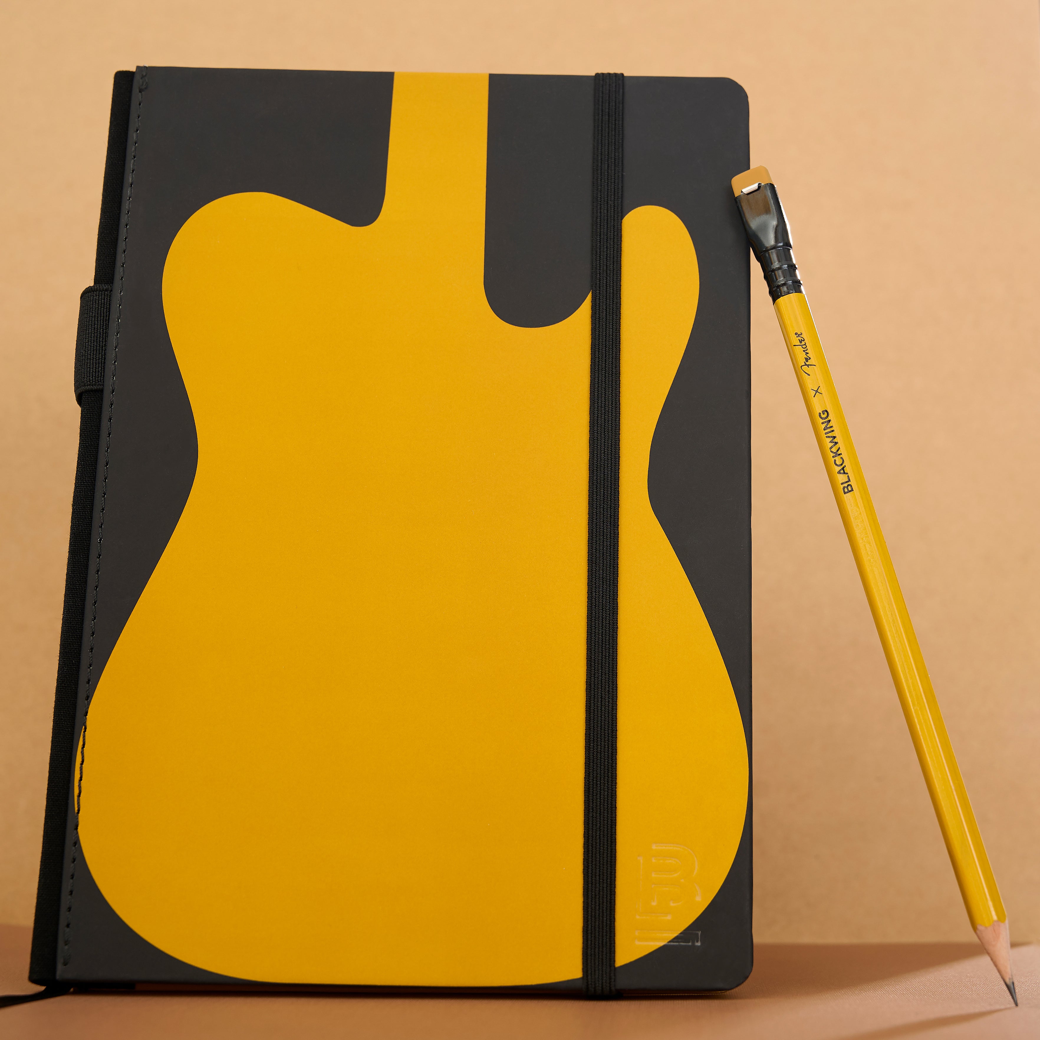 A notebook with a black cover featuring a large yellow guitar-shaped graphic to symbolize the Fender Butterscotch guitar, shown standing upright next to a black and yellow pencil on a beige background.