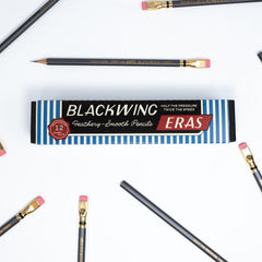 A flat-lay of a Blackwing Eras graphite smooth pencil box on a white background, surrounded by several black pencils with gold ferrules and pink erasers.