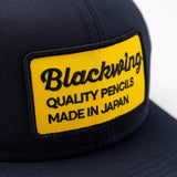 Blackwing trucker hat with yellow "Quality Pencils Made in Japan" embroided logo on front. Navy blue front, white mesh back. Adjustable snapback.