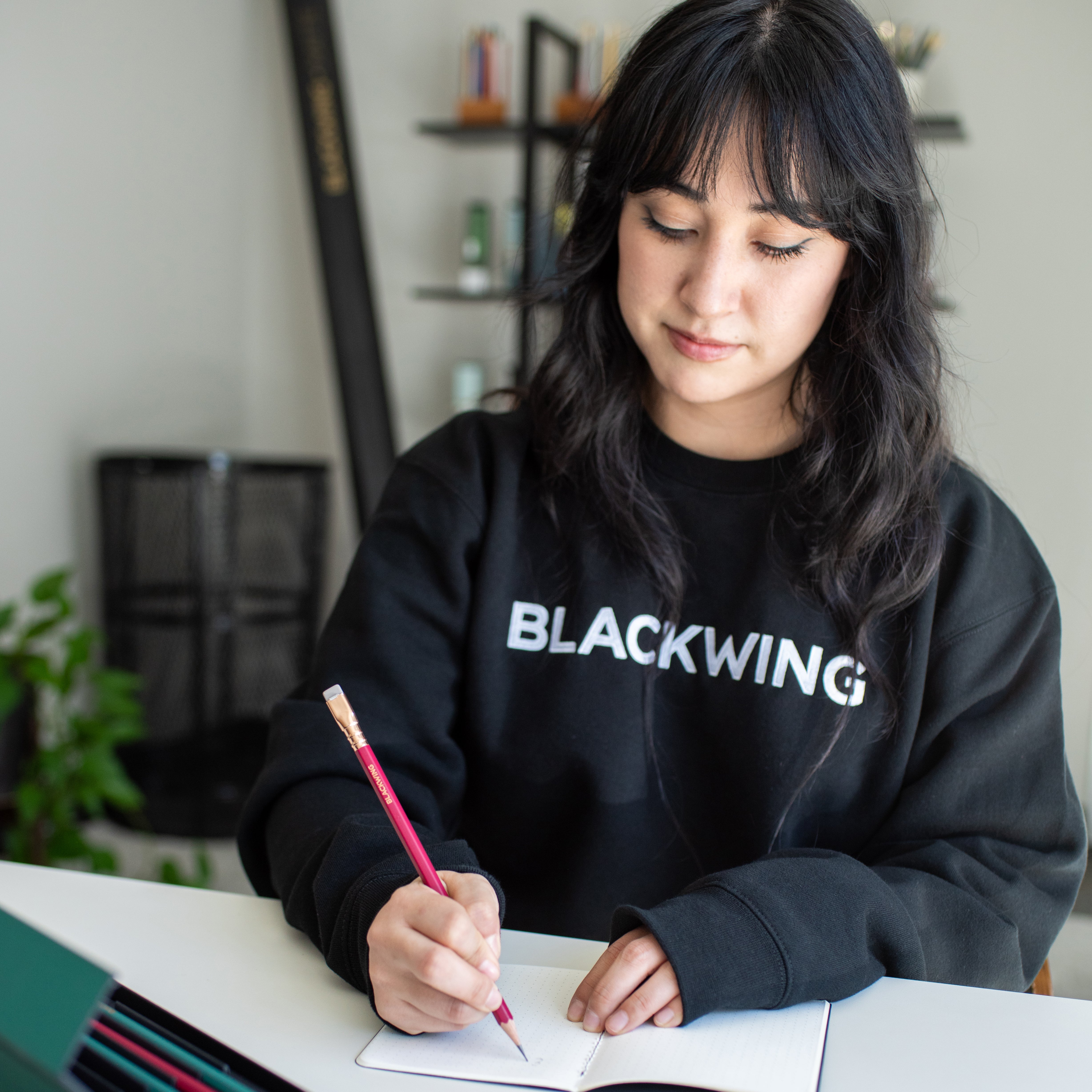 Woman sitting at a desk wearing a black crewneck sweater, writing with a red pencil into a memo book.