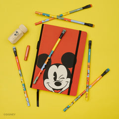 A red Mickey Mouse notebook with a black elastic closure is arranged on a bright yellow background with colorful Disney character pencils, a red point guard, and a pencil sharpener. Disney branding is visible on the corner watermark.