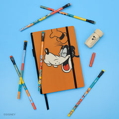 An orange Goofy-themed notebook with a black elastic closure is arranged on a light blue background with matching multicolor pencils, a red point guard, and a cream colored pencil sharpener with black Mickey Mouse face. 