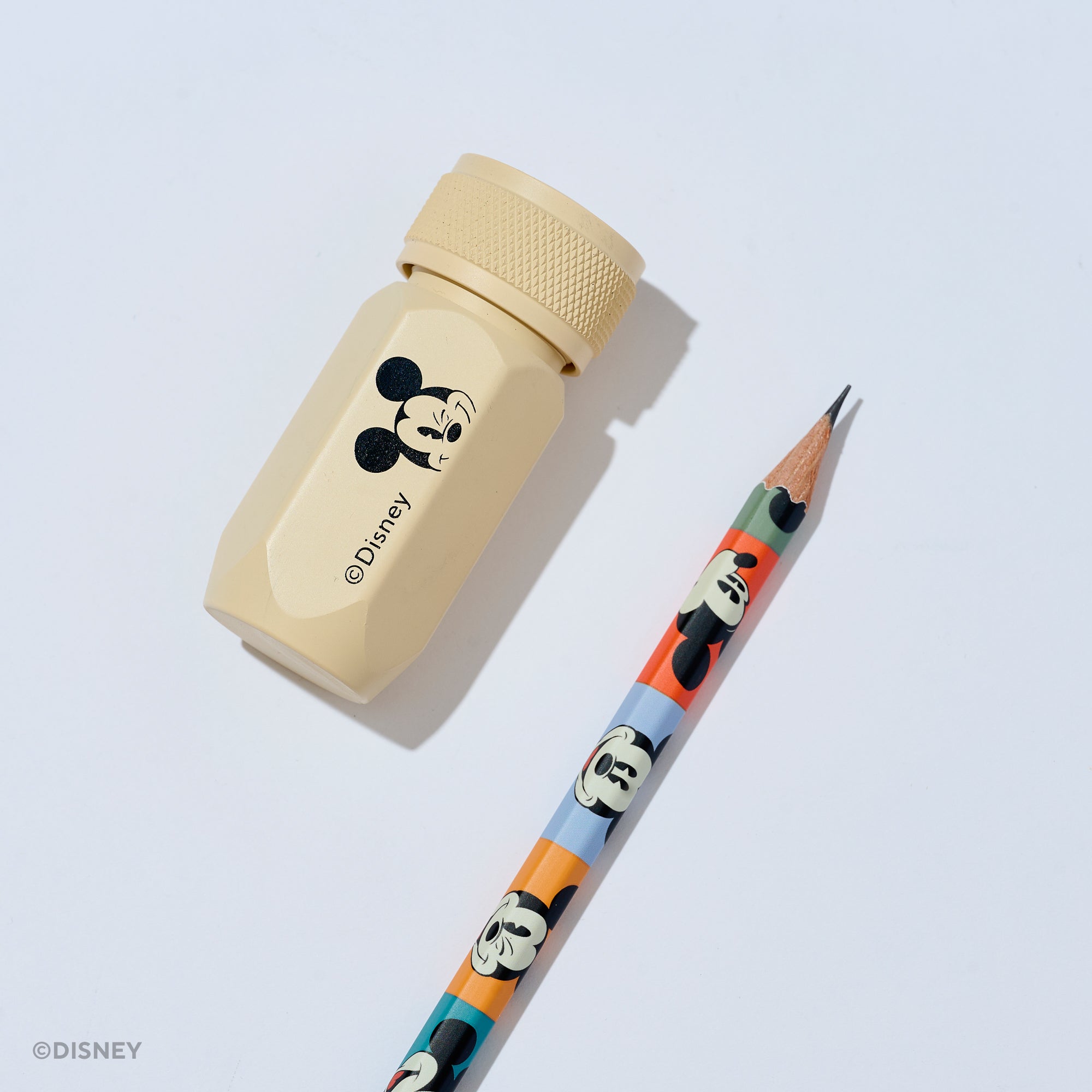 A Disney pencil and pencil sharpener set on a light gray background. The sharpener is a light beige aluminum hexagonal tool with a black Mickey Mouse graphic and the Disney name. The pencil barrel features various Mickey Mouse face graphics in different colorblocks.
