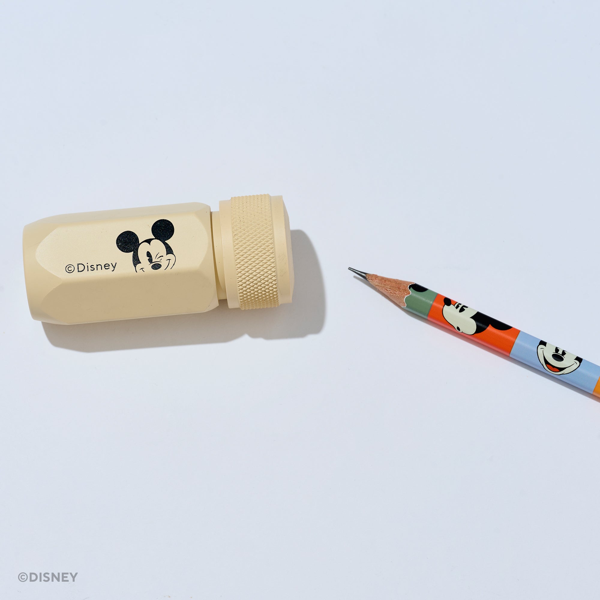 A Disney Mickey Mouse pencil sharpener in a light beige color with a black Mickey graphic and copyright text, shown next to a colorful pencil on a clean white background.