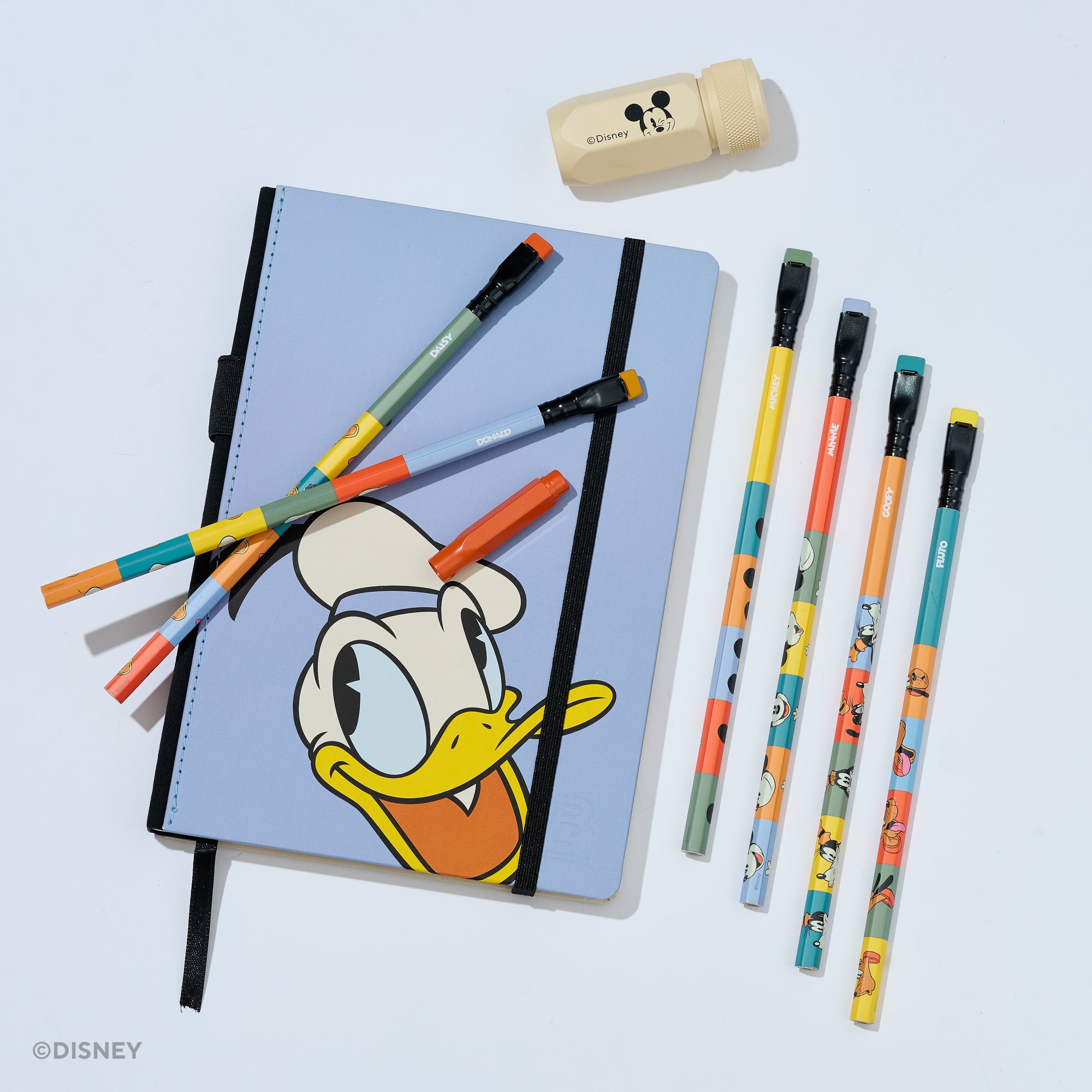 A Disney-themed stationery set on a light gray background, featuring a light blue notebook with Donald Duck artwork, six colorful pencils, a beige Mickey Mouse sharpener, and a red pencil point guard.
