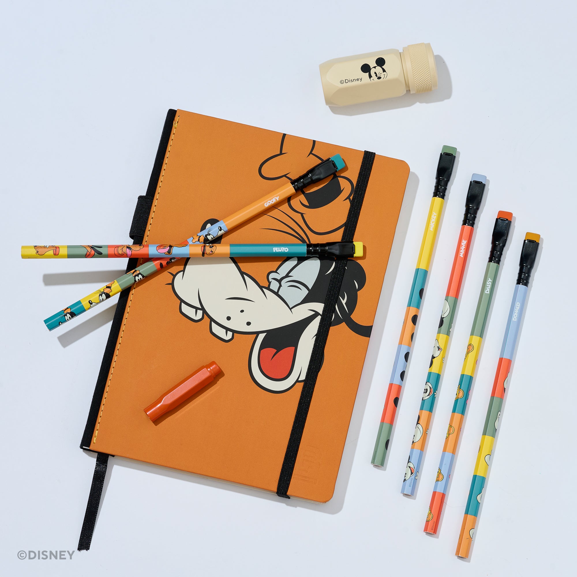 An orange hardcover notebook with a black elastic closure and a Goofy character print, styled with matching pencils, a red point guard, and a cream colored Mickey sharpener on a white background. 