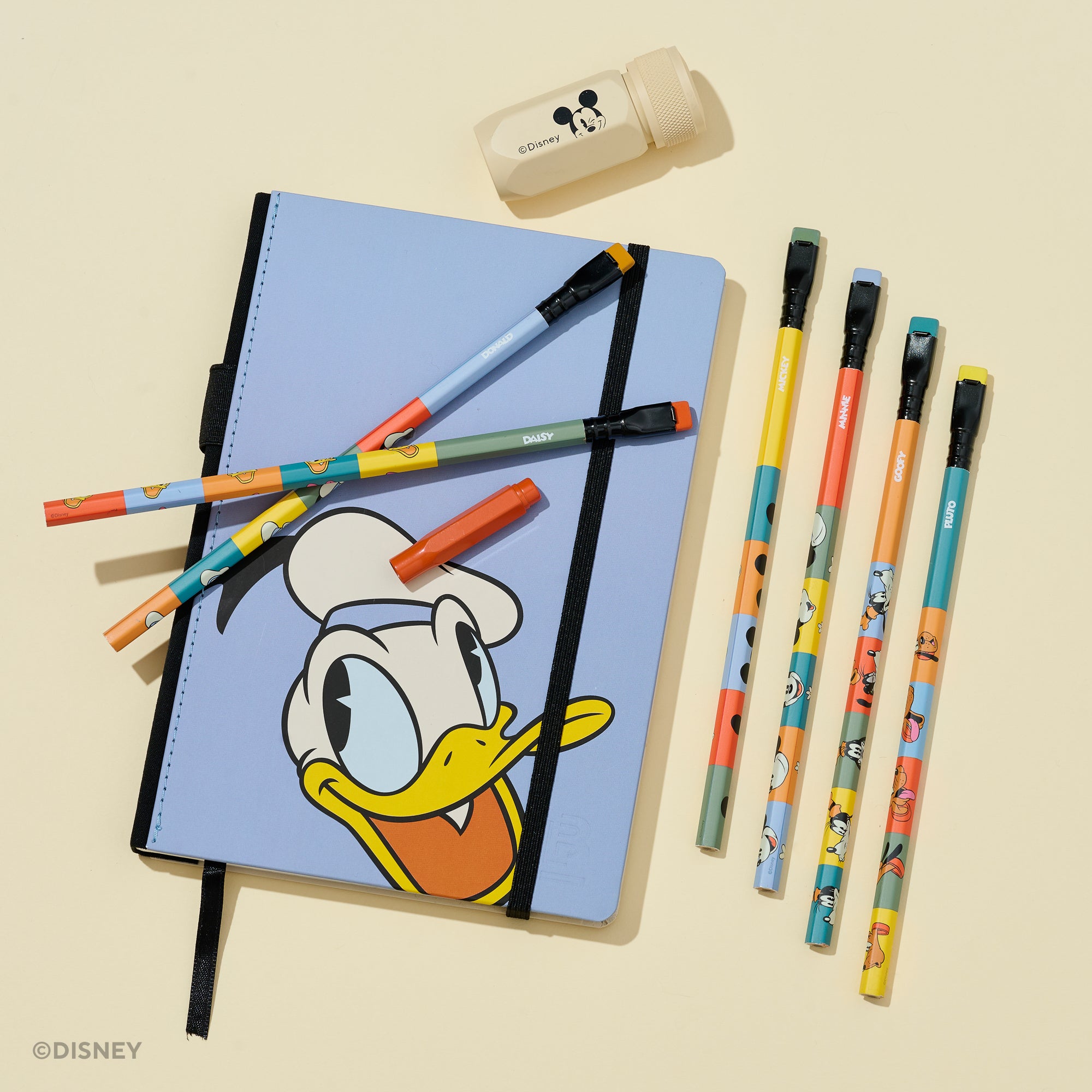 A Disney-themed stationery set on a warm cream background, featuring a light blue notebook with Donald Duck artwork, six colorful pencils, a small red point guard, and a cream pencil sharpener with Mickey Mouse face illustration. 
