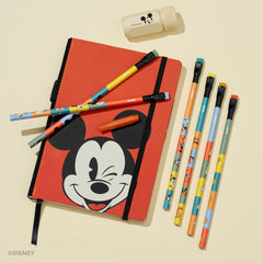 Disney-themed stationery set featuring a red Mickey Mouse notebook with a black elastic closure, six colorful matching pencils, a red point guard, and a small beige sharpener on a light cream background.