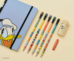 A Disney-themed stationery set arranged on a warm cream background, including a light blue notebook with Donald Duck artwork, matching colorblock pencils, a beige sharpener with Mickey Mouse face, and a red pencil point guard. A Disney watermark appears in the lower left corner.