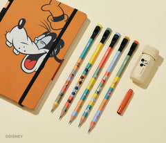 A Disney-themed stationery set arranged on a warm cream background, including an orange notebook with a Goofy illustration, matching pencils, a small red pencil point guard, and a cream sharpener. The Disney watermark appears in the lower-left corner.