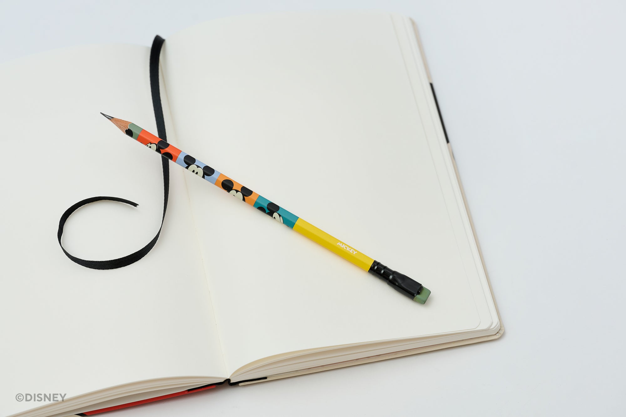 A colorblock Blackwing pencil featuring Mickey Mouse faces rests on an open blank notebook with a black ribbon bookmark, shot on a clean white background.