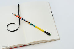 A colorblock Blackwing pencil featuring Mickey Mouse faces rests on an open blank notebook with a black ribbon bookmark, shot on a clean white background.
