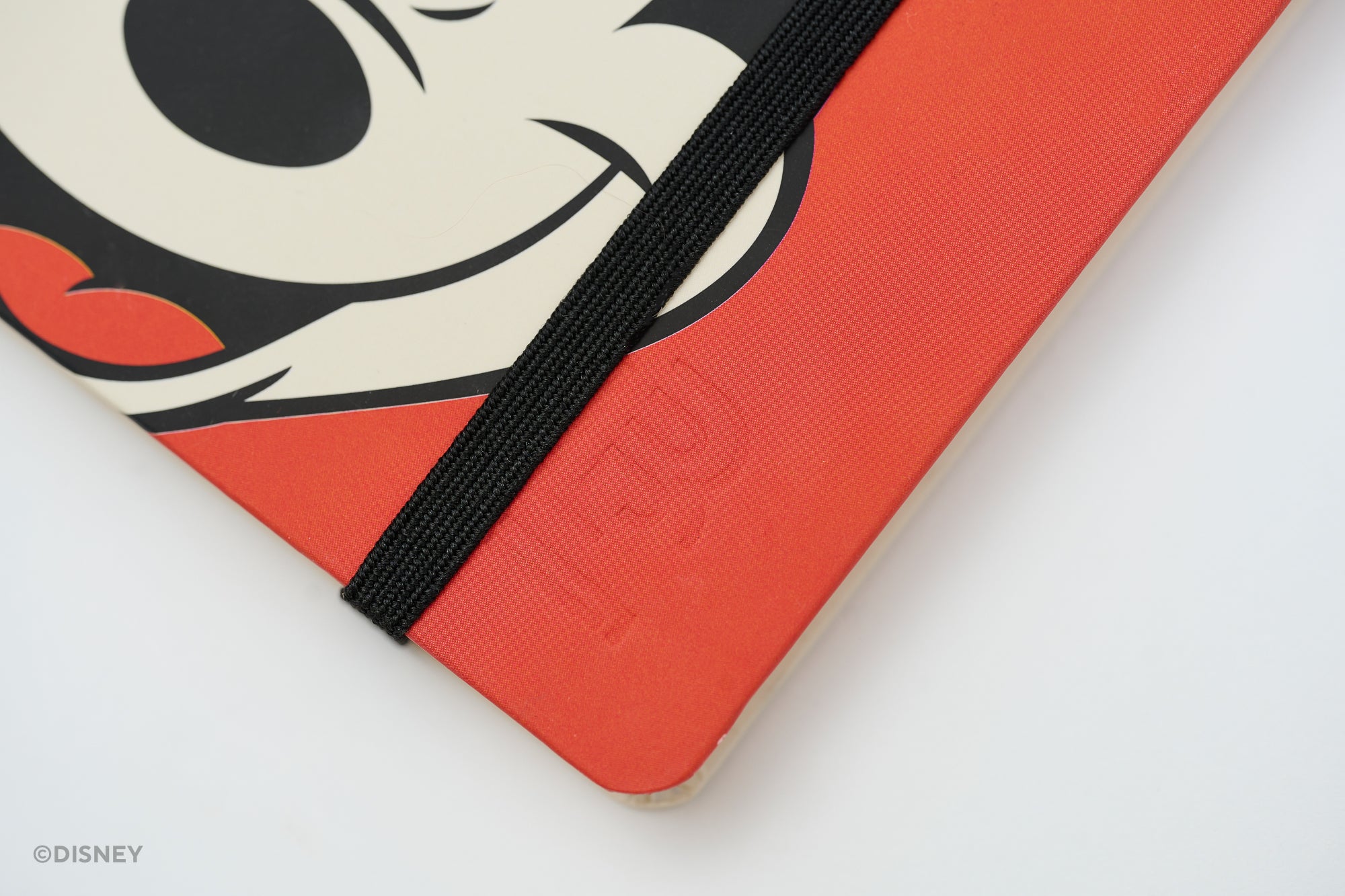 A close-up of a red notebook with Mickey Mouse's face with a black elastic closure band. The Disney watermark is visible in the bottom-left corner.