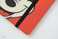 A close-up of a red notebook with Mickey Mouse's face with a black elastic closure band. The Disney watermark is visible in the bottom-left corner.