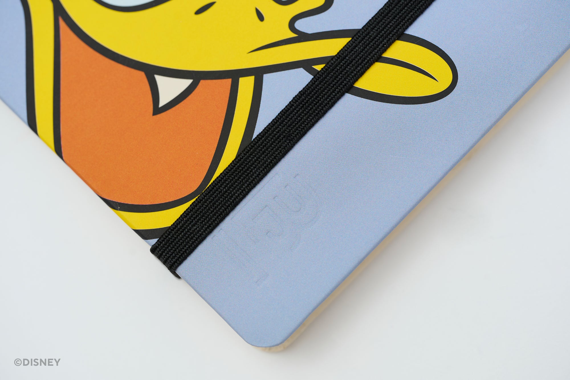 A close-up of a light blue Disney Blackwing notebook with a black elastic closure band and a cartoon Donald Duck face printed on the cover. A Disney watermark is visible in the bottom-left corner.