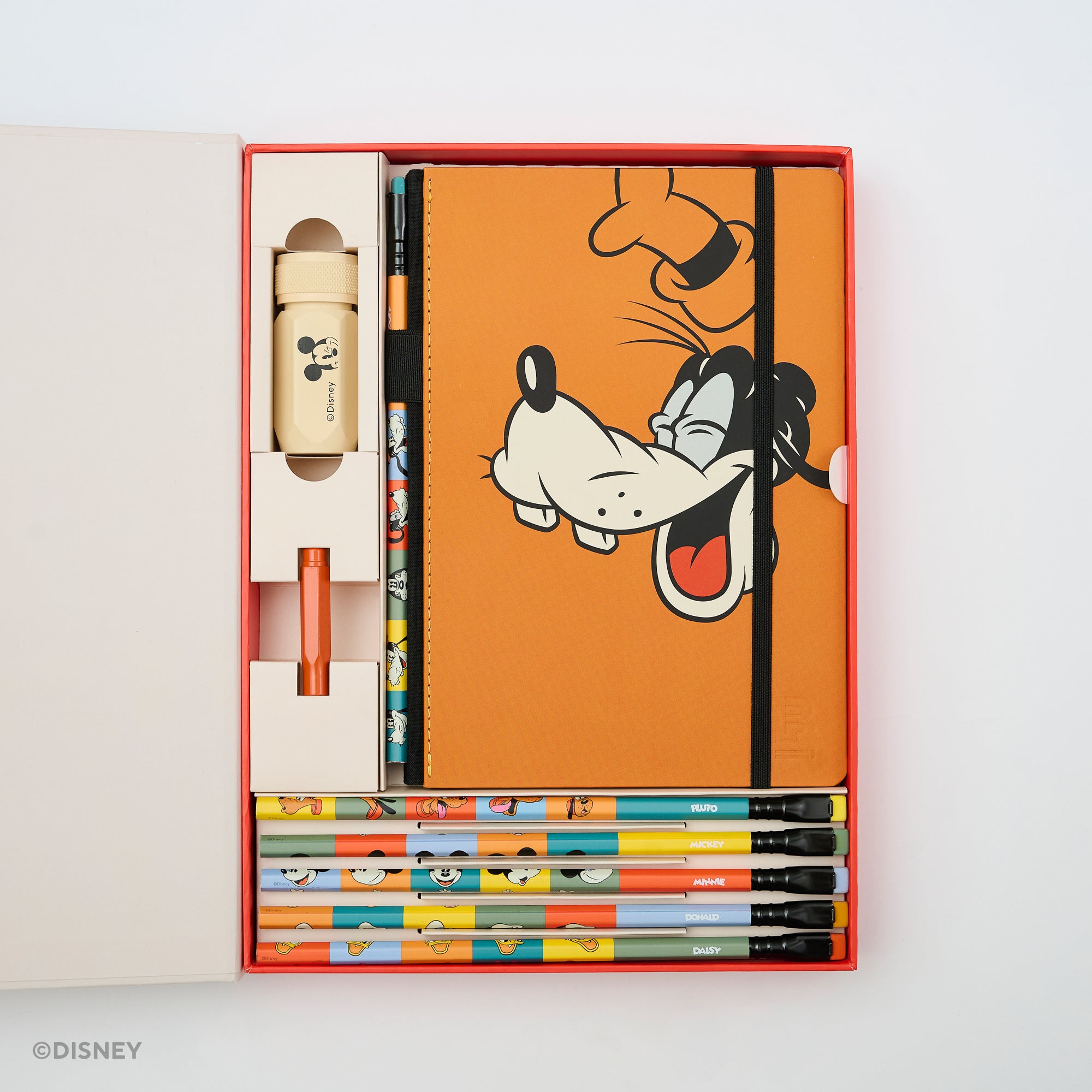 A Disney-themed stationery gift set displayed inside an open red and white box on a white background. The set includes an orange notebook with a Goofy illustration, several colorful pencils, a cream Mickey Mouse pencil sharpener, a a red point guard.