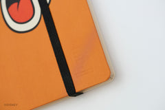 Close-up of an orange hardcover notebook with a black elastic closure band and a partial Goofy face. A faint embossed Blackwing logo is visible on the cover, with a small Disney watermark in the lower-left corner.