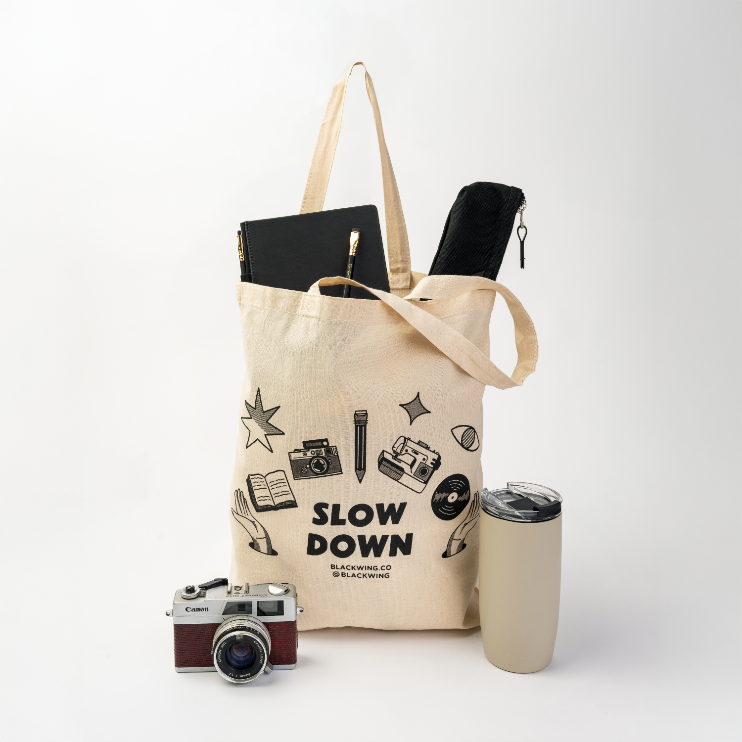 canvas tote that says slow down in black with graphics of camera, pencil, sewing machine, record, and book