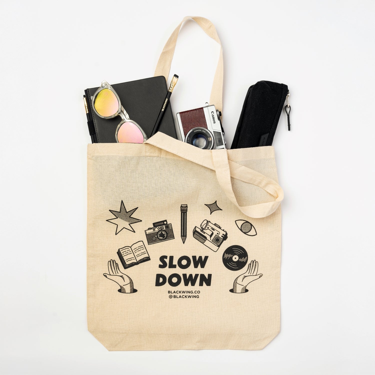 canvas tote that says slow down in black with graphics of camera, pencil, sewing machine, record, and book