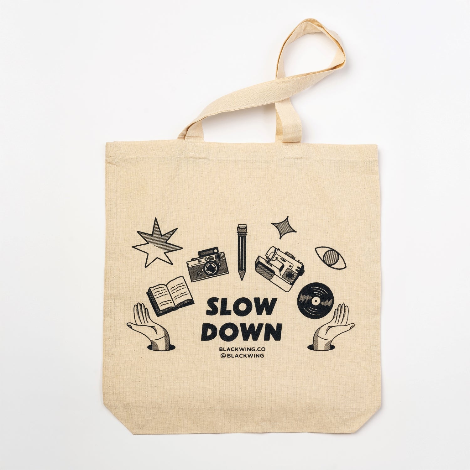 canvas tote that says slow down in black with graphics of camera, pencil, sewing machine, record, and book