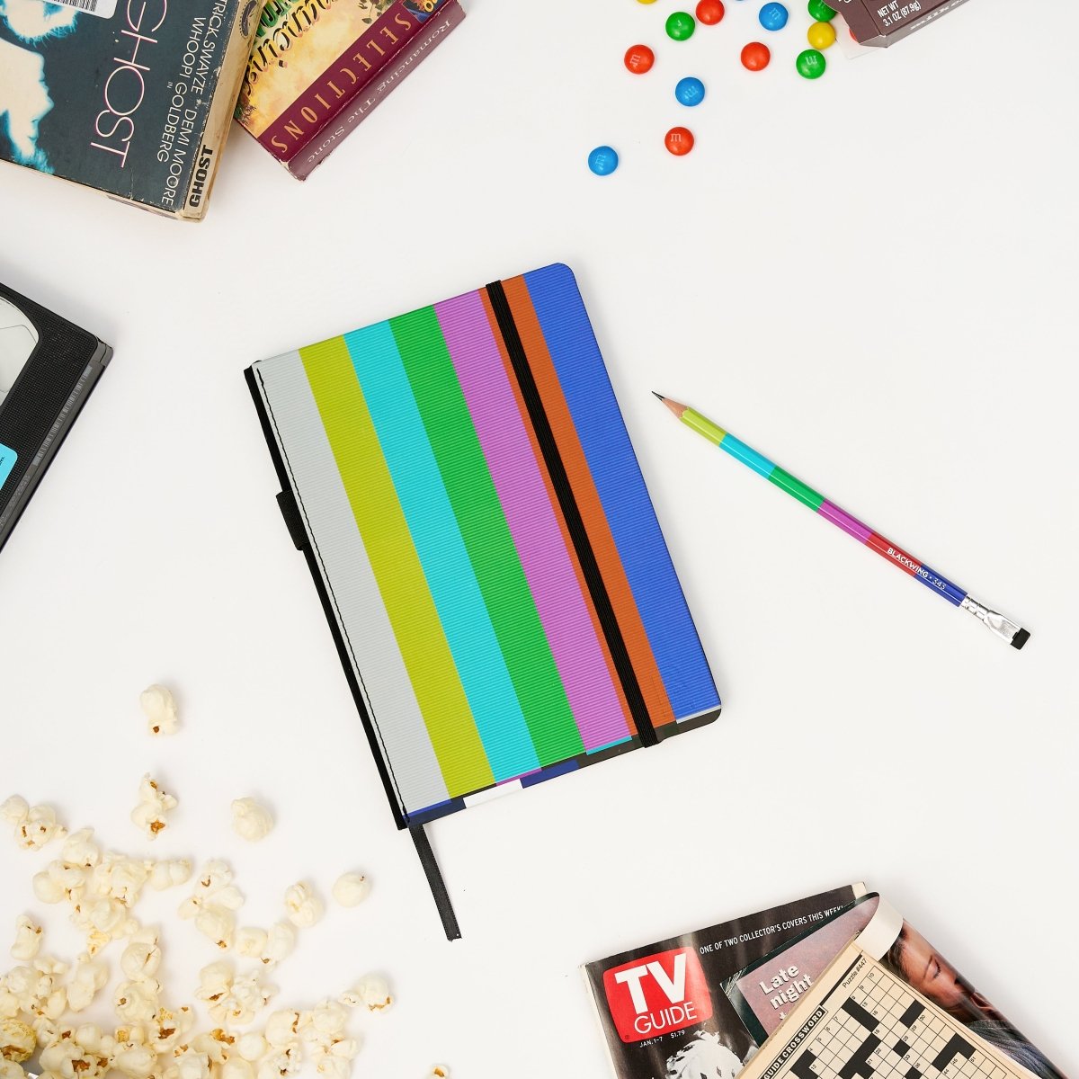 Colorful striped hardcover notebook with a matching pencil laying next to popcorn, m&ms, and TV guide on a white surface