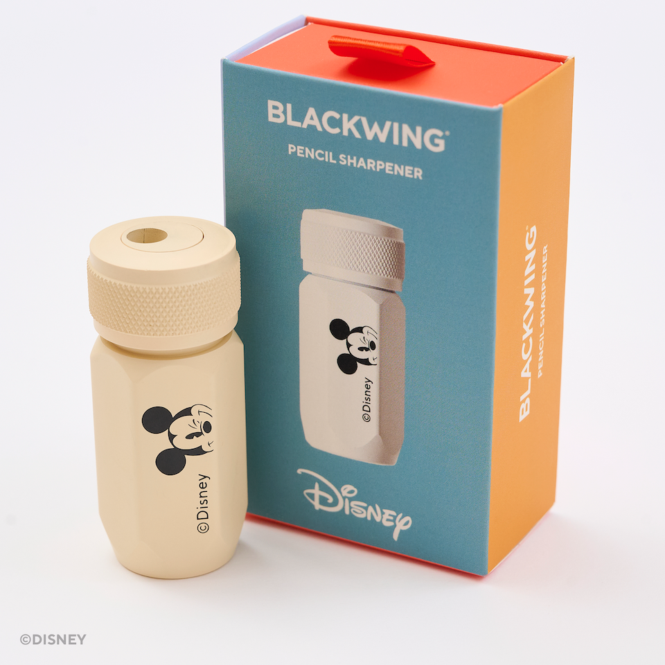 A Blackwing Disney Pencil Sharpener shown next to its retail packaging box. The sharpener is a light cream color with a black Mickey Mouse graphic and a matching textured cap.